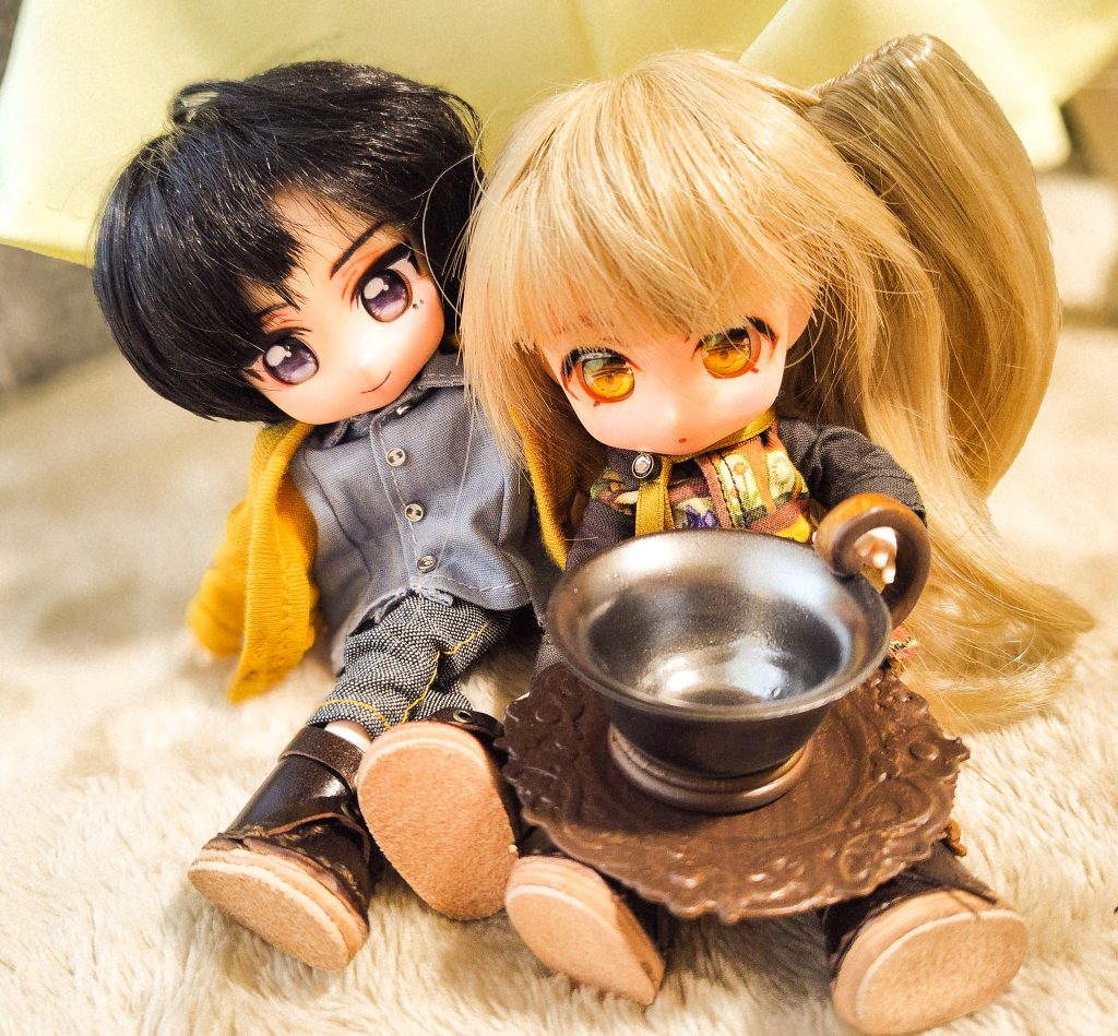 小さなドールが大きなティーカップをひざにのせている。もう一人のドールがそれを後ろからのぞき込んでいる。